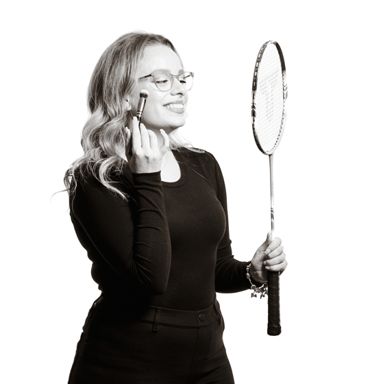 Sandra grinst während sie so tut als würde sie den Badmington-Schläger als Spiegel benutzen um sich zu schminken.