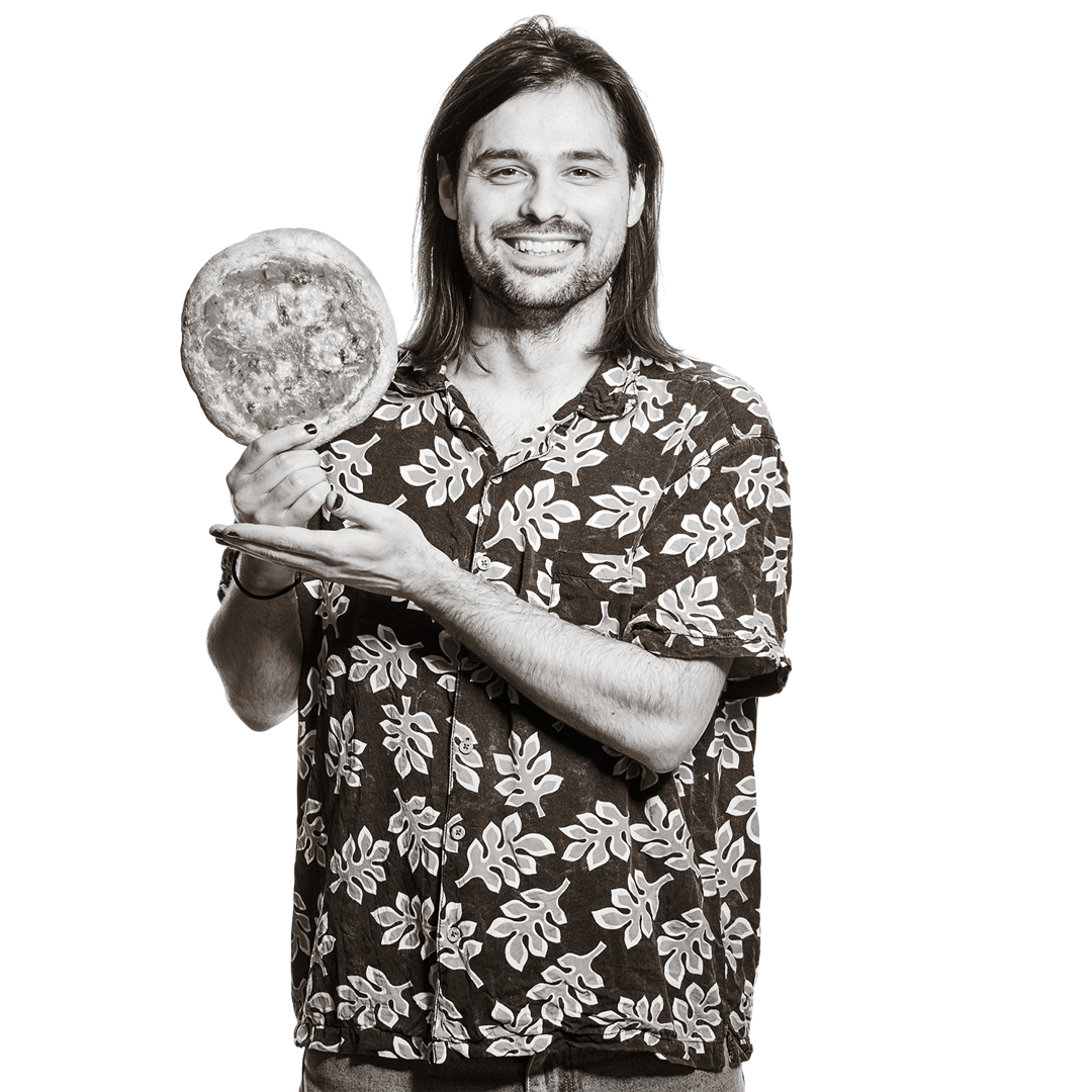 Portrait von Nicolas. Er hält breit lächelnd eine kleine Pizza in die Höhe. 