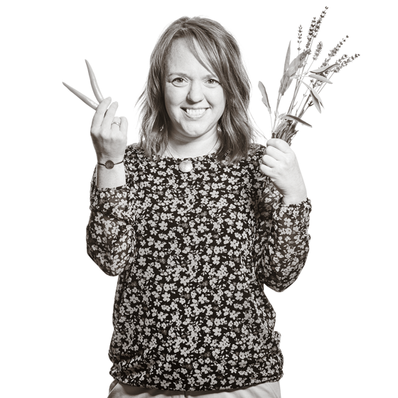 Claudia hält in einer Hand zwei Karotten hoch, in der anderen ein Büschel Kräuter.
