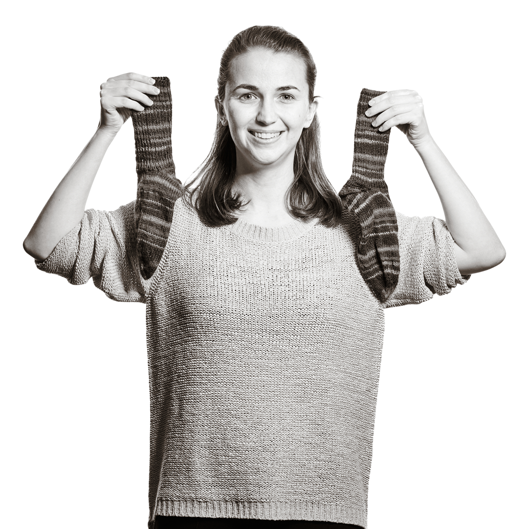 Portrait von Verena. Sie hält zwei Stricknadeln, an denen schon ein Stück fertiger Schal zu sehen ist.